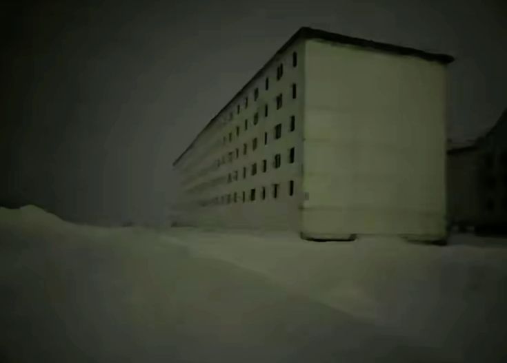 Local a noite escuro e vazio, com neve e um prédio de apartementos coberto por neblina